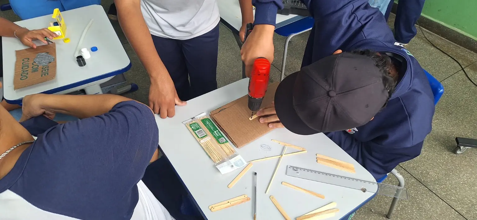 Fotografia do processo de fabricação da base. Um estudante com boné perfura cuidadosamente uma peça de papelão apoiada na mesa, enquanto colegas acompanham. Ao redor há palitos de sorvete, régua, cola, barbante, tampas e tintas. O boné e o ângulo da câmera ocultam o rosto; os demais rostos não aparecem.