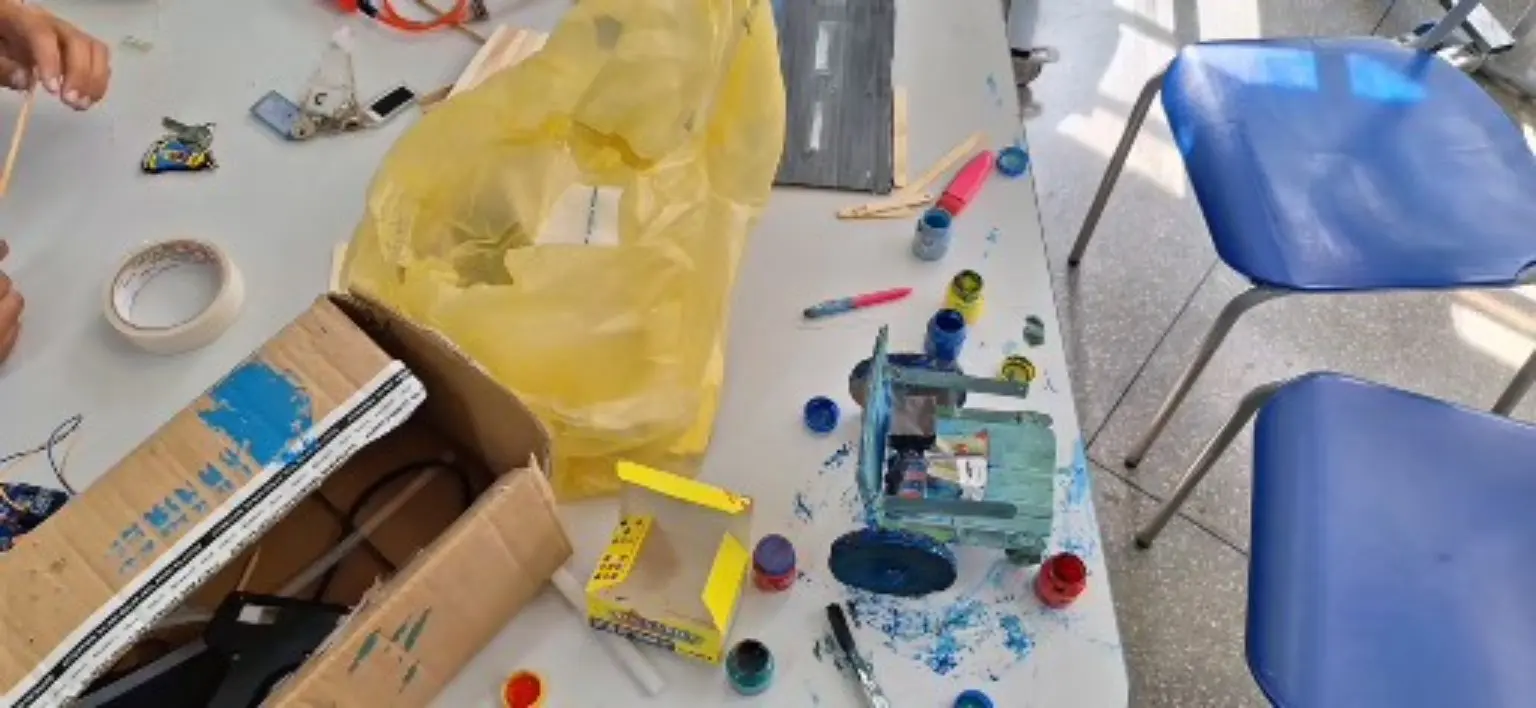 Vista superior de uma mesa durante a construção. À direita está a cadeira artesanal azul, já com encosto, assento e rodas grandes. Ao centro aparecem tintas azul e vermelha, pincel, palitos e uma faixa de papelão pintada como pista. À esquerda há uma caixa com materiais. Apenas mãos e braços entram no enquadramento.