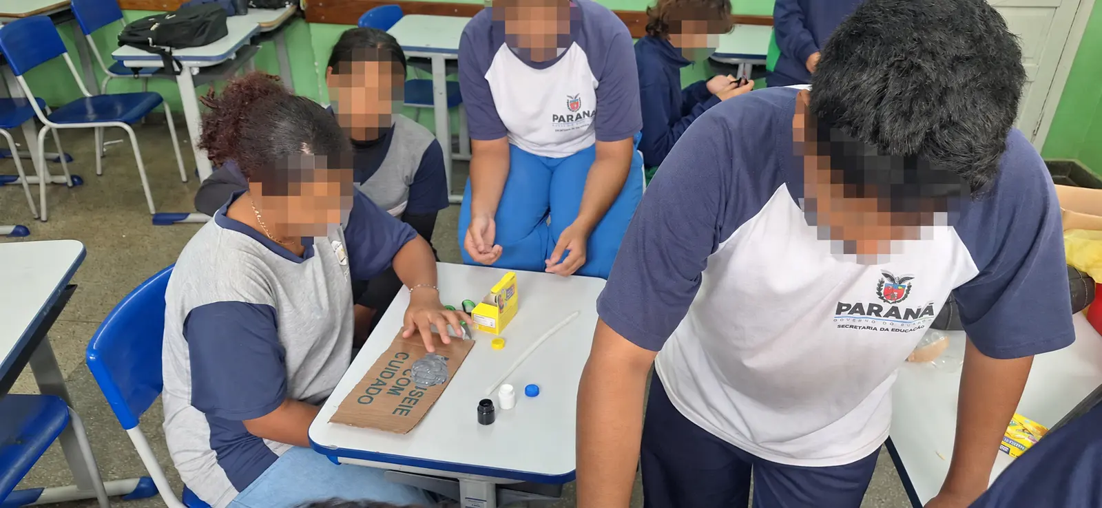 Grupo de estudantes reunido em torno de uma mesa de sala de aula durante a confecção de uma placa de papelão. Tintas, tampas, cola e outros materiais estão espalhados sobre a mesa. Todos os rostos visíveis receberam desfoque pixelado forte para preservar a identidade dos estudantes.