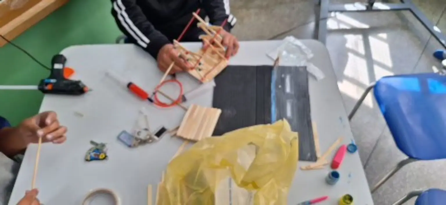 Mesa de sala de aula vista de cima. Estudantes montam uma estrutura articulada com palitos de sorvete sobre a futura pista de papelão. Uma pistola de cola quente, tesoura, régua, tintas e conjuntos de palitos estão distribuídos pela mesa. Os rostos ficam fora do enquadramento.