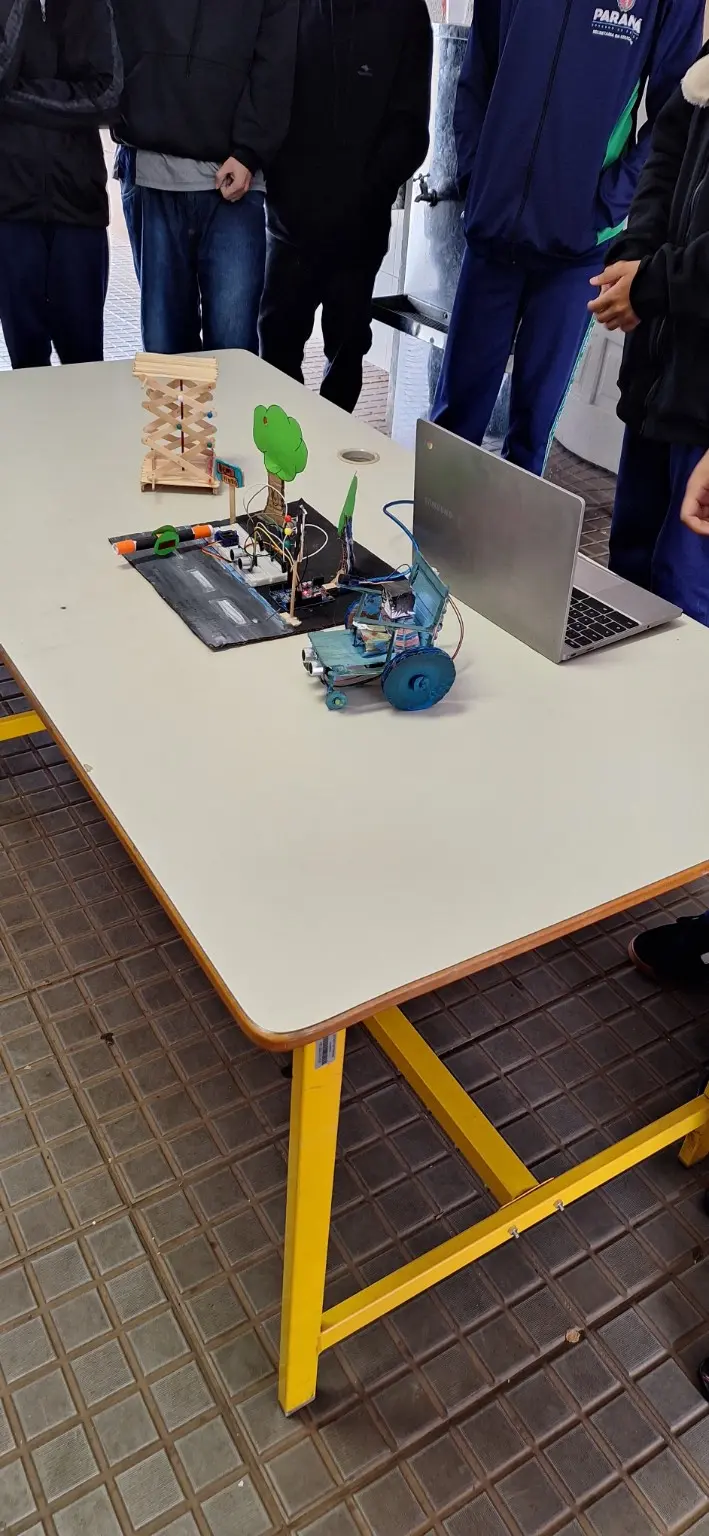 Fotografia vertical do trabalho final sobre uma mesa escolar. A maquete reúne, da esquerda para a direita, o elevador pantográfico de palitos, uma pista preta com cancela, árvore, semáforo, sensores, Arduino e protoboard, e uma cadeira artesanal azul com grandes rodas. Um notebook aberto está ao lado da montagem. Estudantes permanecem em volta da mesa, enquadrados do pescoço para baixo, sem rostos identificáveis.