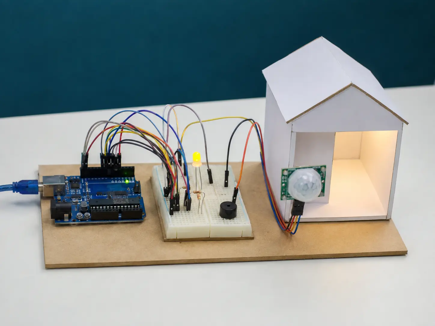 Protótipo inteiro de sensor PIR sobre base de MDF, com Arduino, protoboard, fios, LED amarelo, buzzer e uma pequena casa de papel iluminada; não há pessoas.